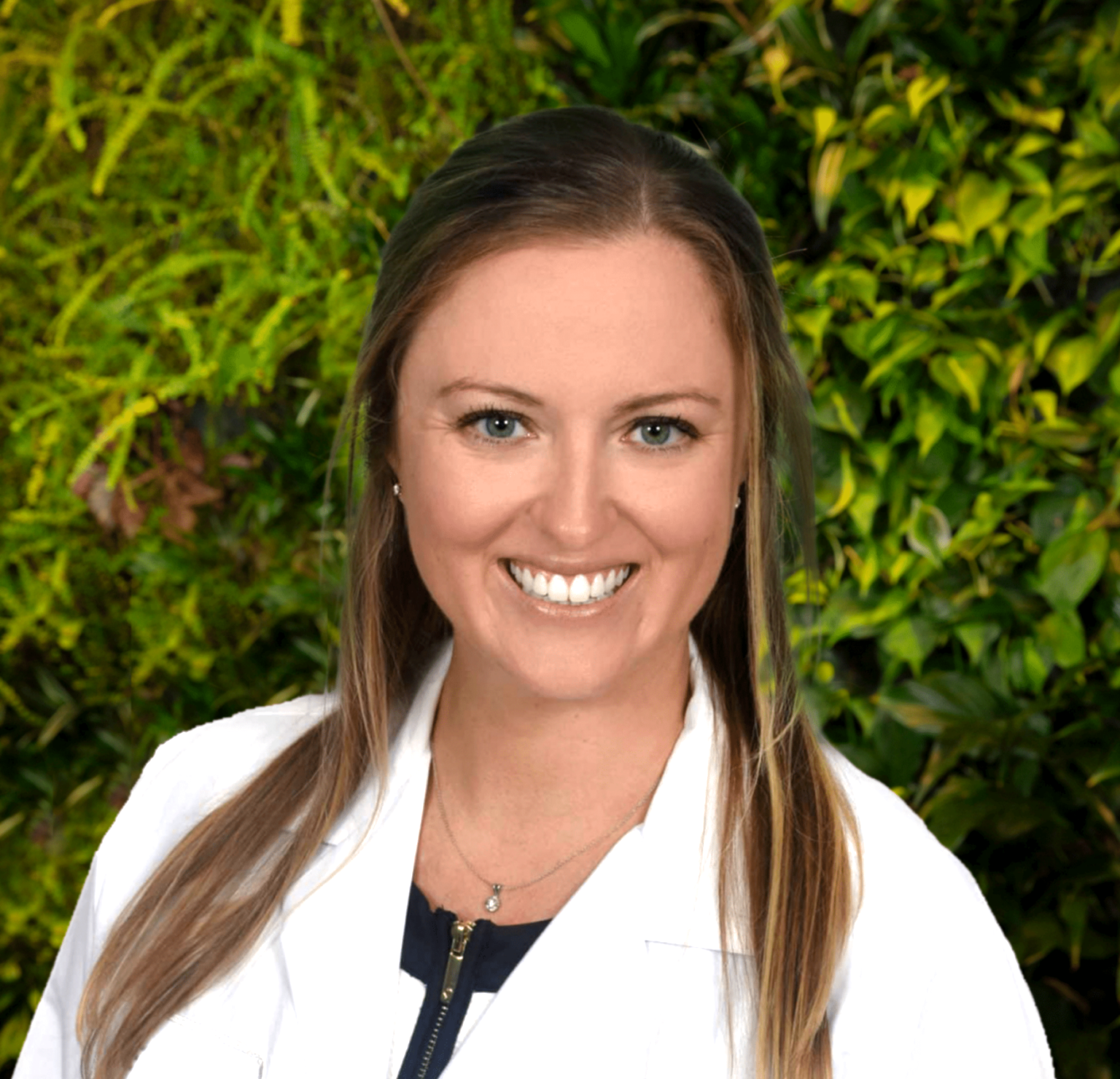 Smiling person with long hair in white coat, lush green foliage background.