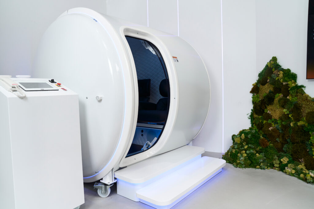 Cylindrical hyperbaric chamber with open door, control panel, in modern room with greenery.