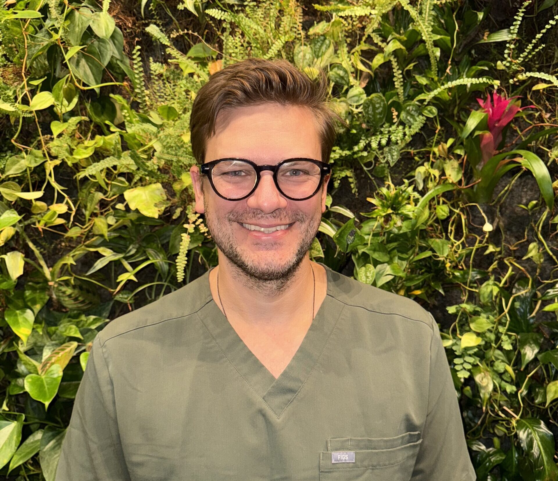 Person in glasses with green scrubs smiling, standing before plant-filled wall with pink flower.