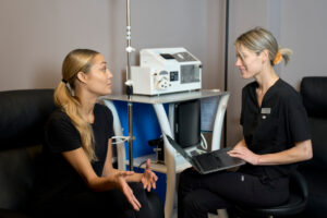 Healthcare professional with laptop consulting a person in a clinical setting with medical equipment.