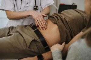 Healthcare professional examining patient's abdomen with another person present holding an arm.