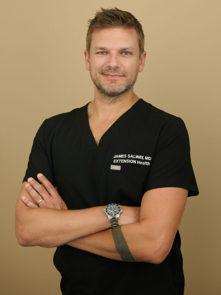 Man in black medical scrubs with James Salway MD, Extension Health, embroidered, arms crossed.