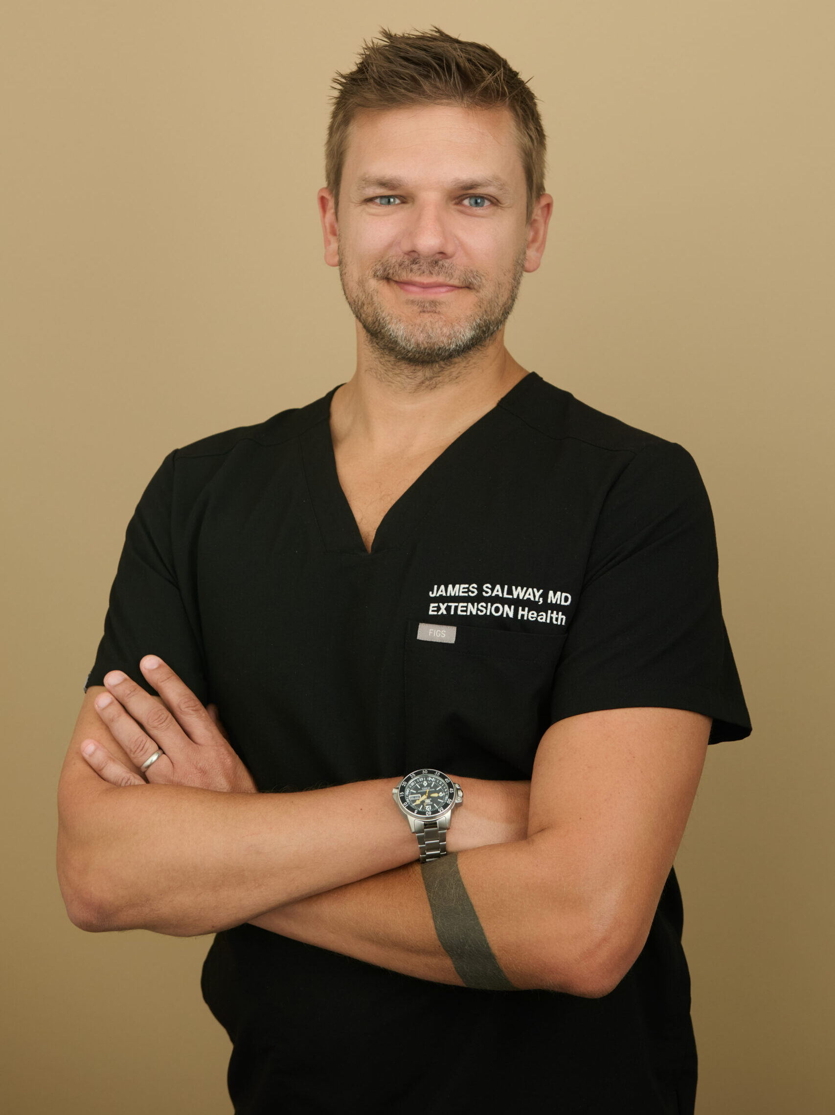 Man in black medical scrubs with James Salway MD, Extension Health, embroidered, arms crossed.