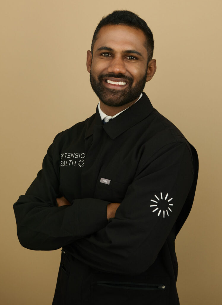 Man in black Extension Health jacket with circular logo on sleeve standing with arms crossed on beige background