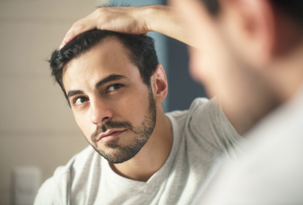 Man in light shirt, touching hair, looking thoughtfully in mirror with facial hair.