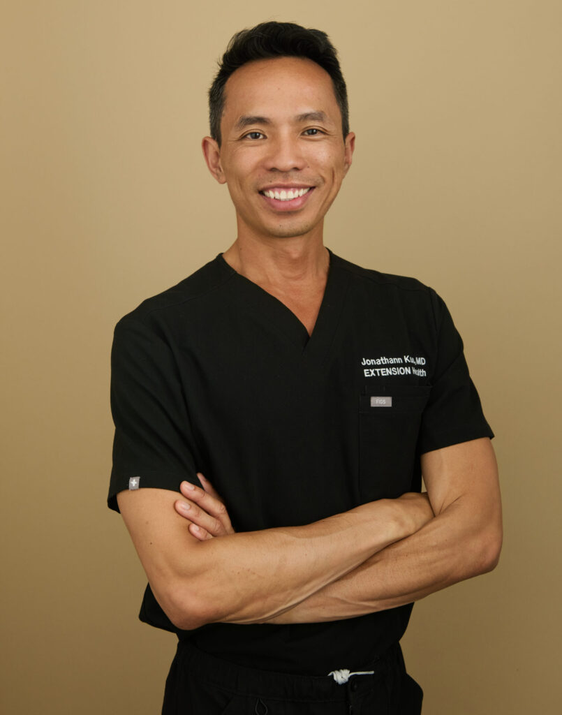 Man in black medical scrubs with Jonathan Ku MD Extension Health embroidery standing with arms crossed.