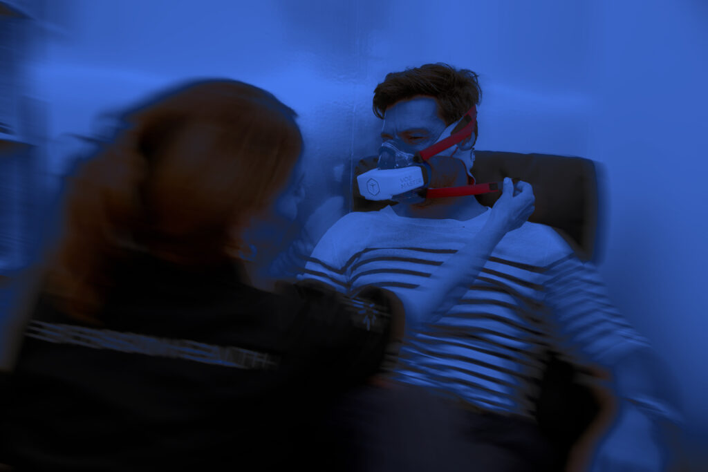 Person seated wearing a striped shirt with VO2 Master respiratory mask adjusted by another individual under blue light.