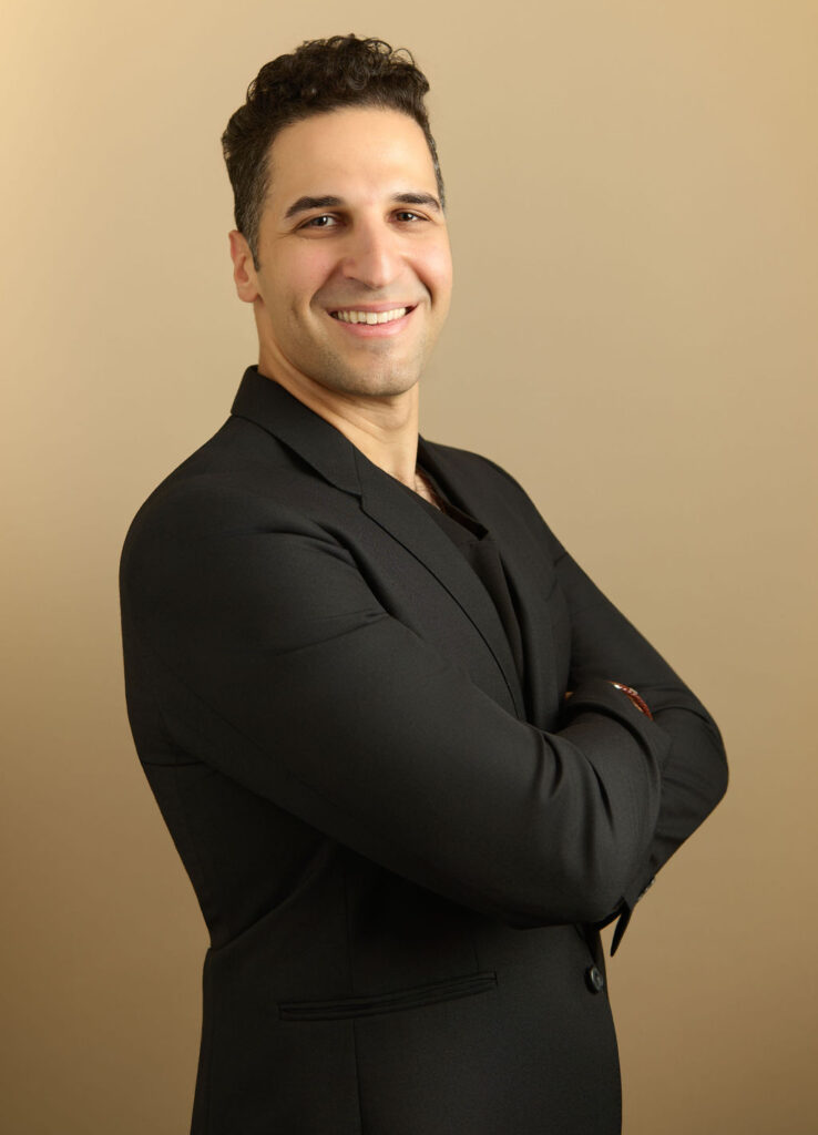Man in a black blazer standing with arms crossed and smiling against a beige background
