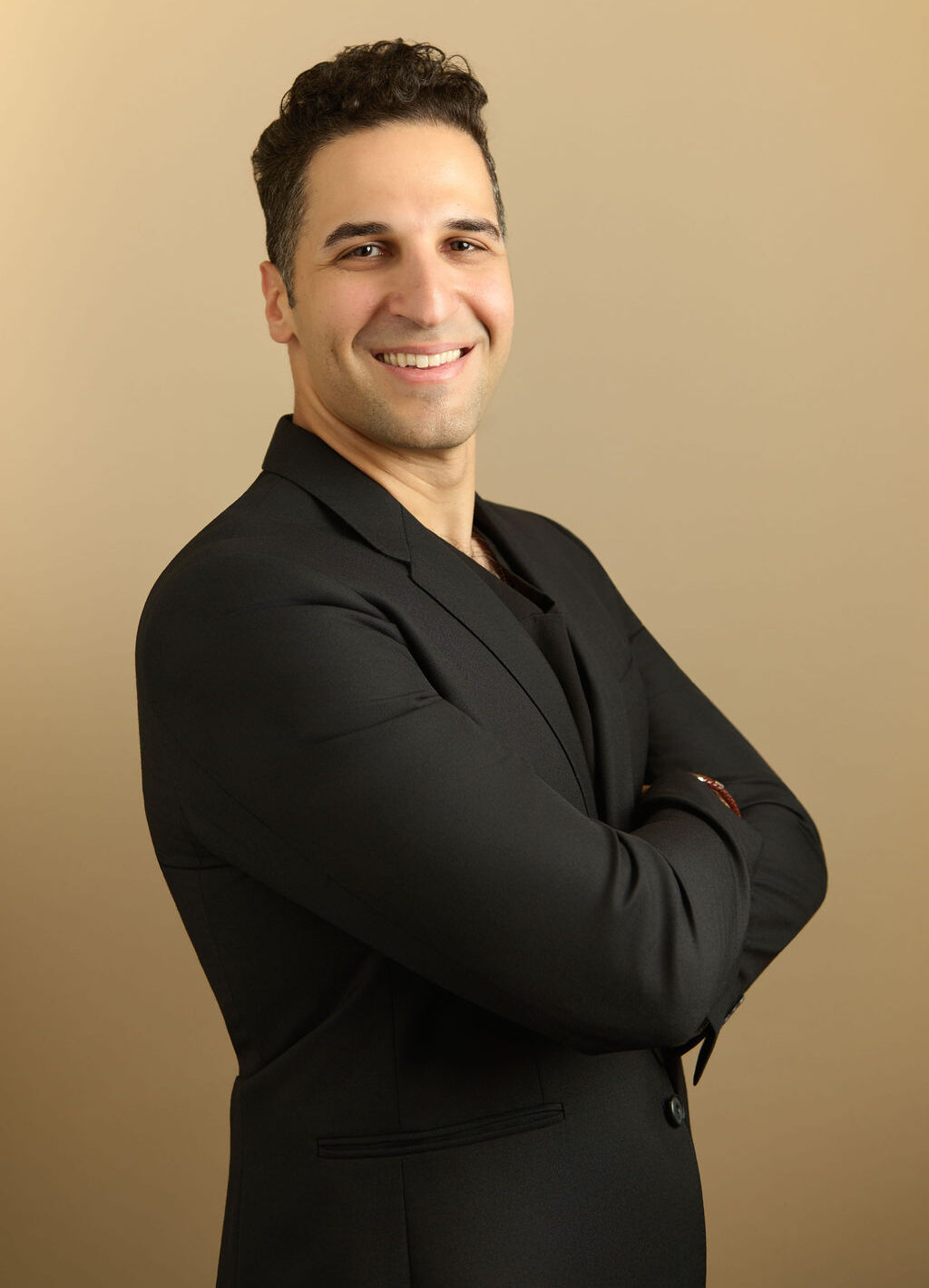 Man in a black blazer standing with arms crossed and smiling against a beige background
