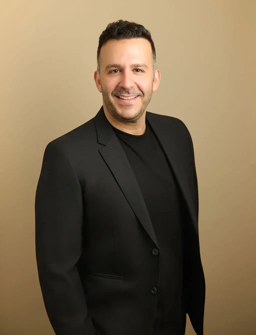 Man with short dark hair wearing a black blazer and shirt smiling against beige background
