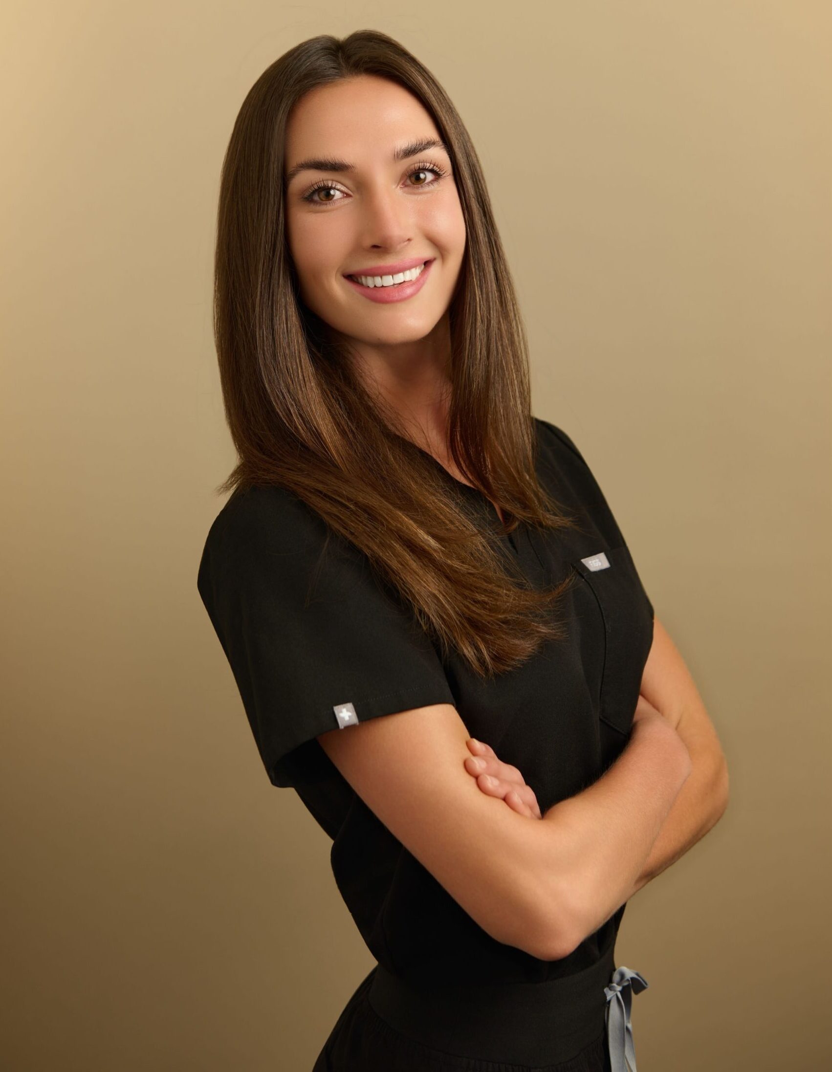 Young woman with long brown hair wearing black scrubs standing with arms crossed.