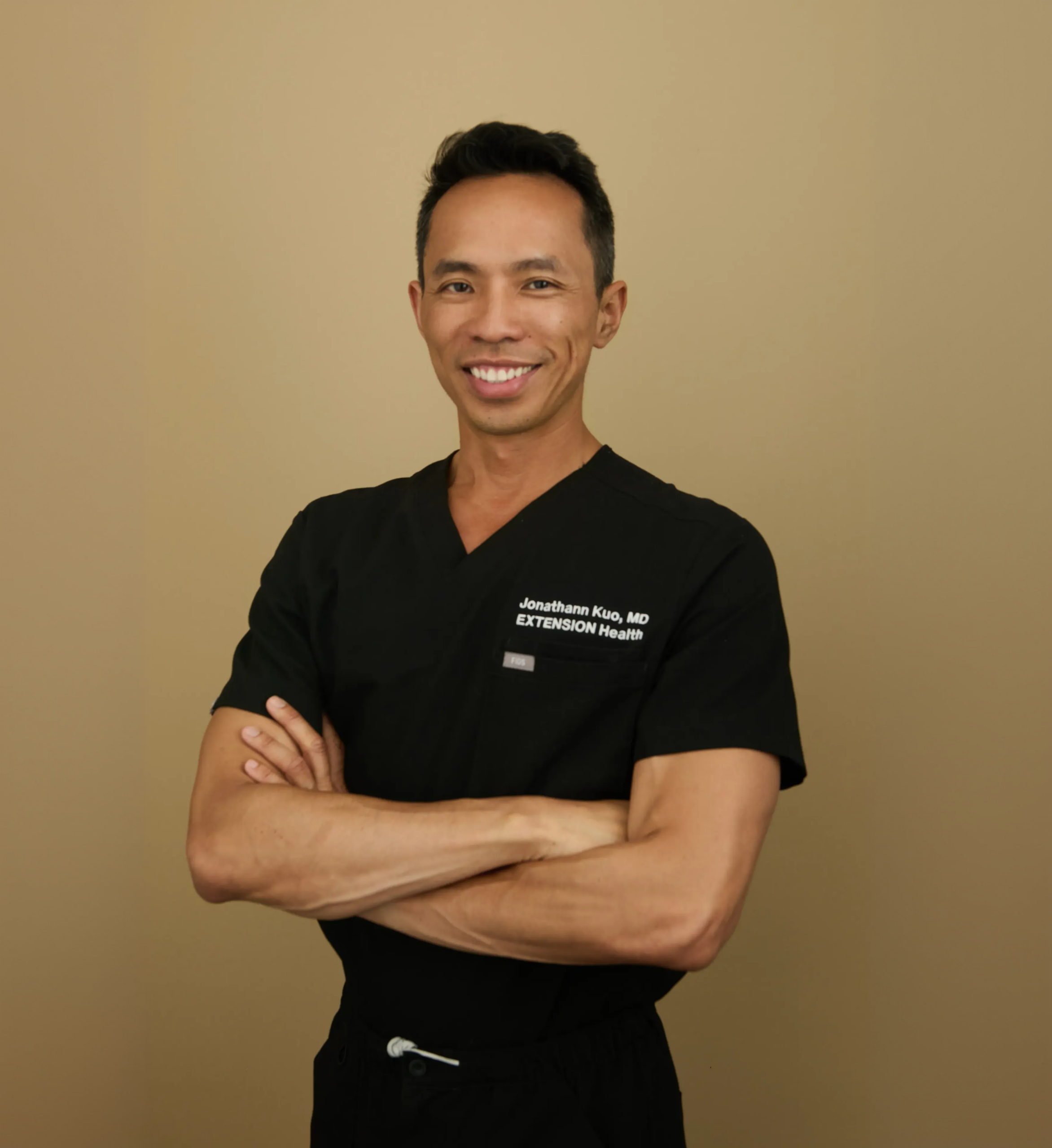 Male doctor in black scrubs with name tag standing with arms crossed on beige background
