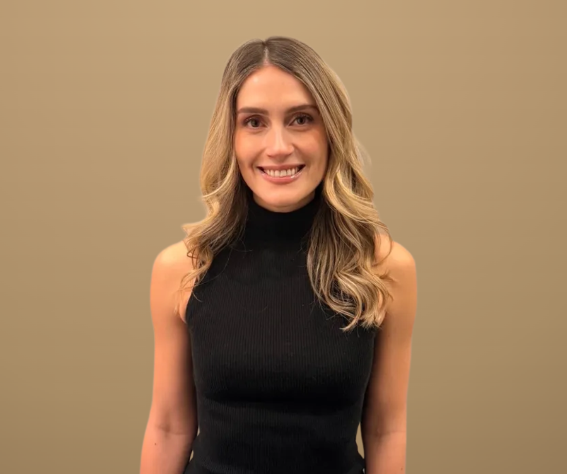 Young woman with long blonde hair wearing a sleeveless black turtleneck and pants, standing against a beige background.