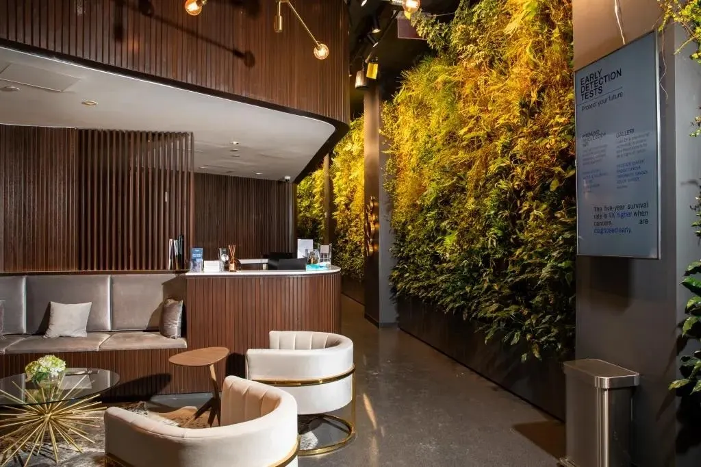 Modern hotel lobby with dark wood, curved reception, gray sofas, white/gold chairs, and a green vertical garden.